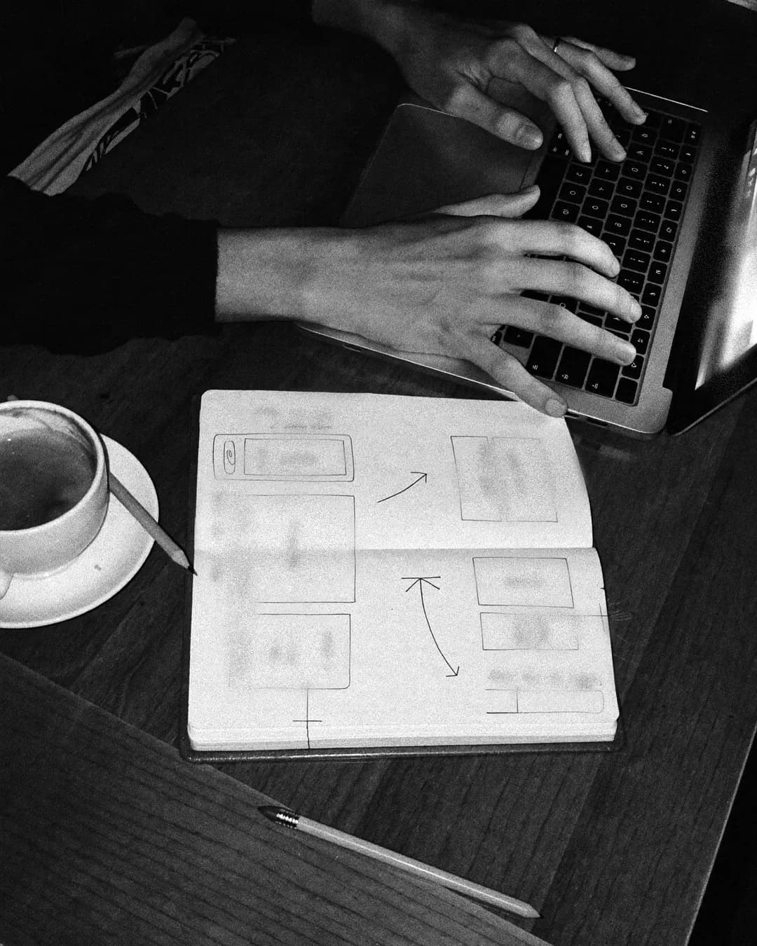 Hands on a laptop next to a notebook with sketches of a web page